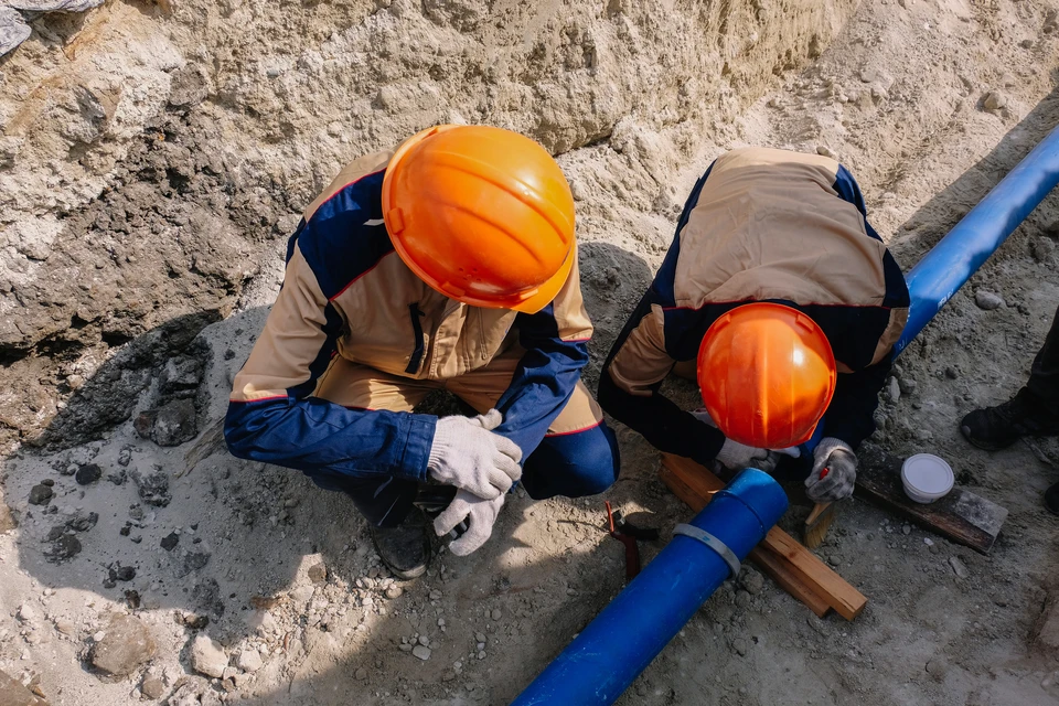 Deux ouvriers équipés de casques de sécurité orange travaillent à genoux sur un chantier extérieur, effectuant des opérations de maintenance ou réparation sur une canalisation bleue enterrée dans le sol sablonneux